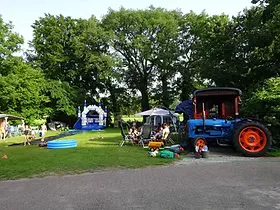 Kampeerveld met een blauwe trekker, een springkussen en kampeerders onder bomen bij Camping Achter 't Koeienpad